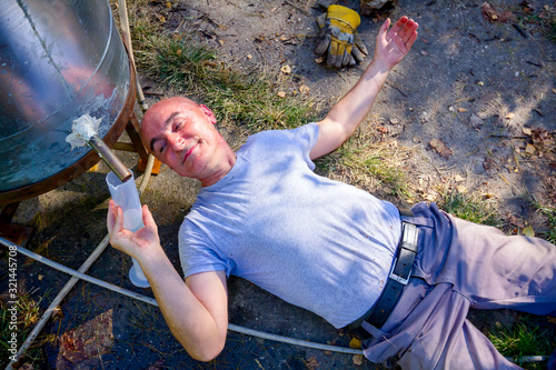 Man lies beneath condenser drinking freshly distillated liquor, alcohol photo