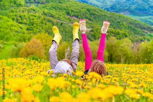 Fototapeta A boy and a girl lie on a meadow of dandelions in the grass in the mountains with their legs up. Happy children have a rest in mountains. Happiness and Carefree