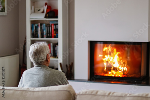 Old thoughtful gray haired man sitting on sofa in front of fireplace at home watching fire. Senior pensioner lifestyle. Loneliness.