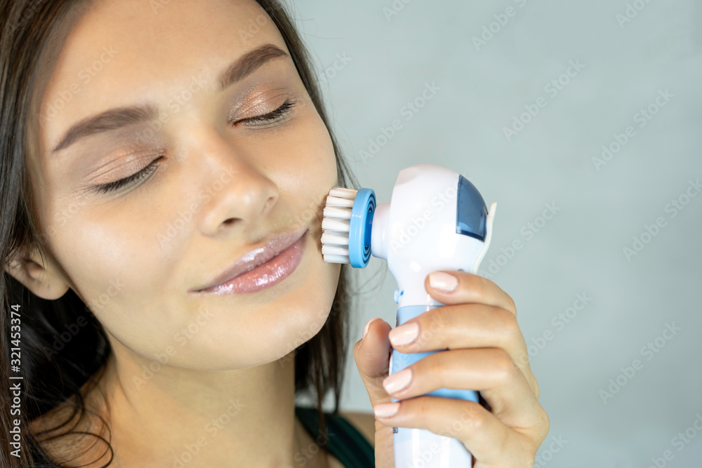 Young pretty woman doing skin care procedure. Cleaning face with machine on grey background copy space Young pretty woman doing skin care procedure. Cleaning face with machine on grey background copy space