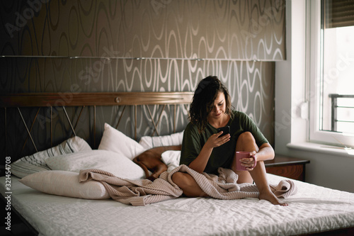 Girl with dog in bed. Woman drinking coffee and using phone. 