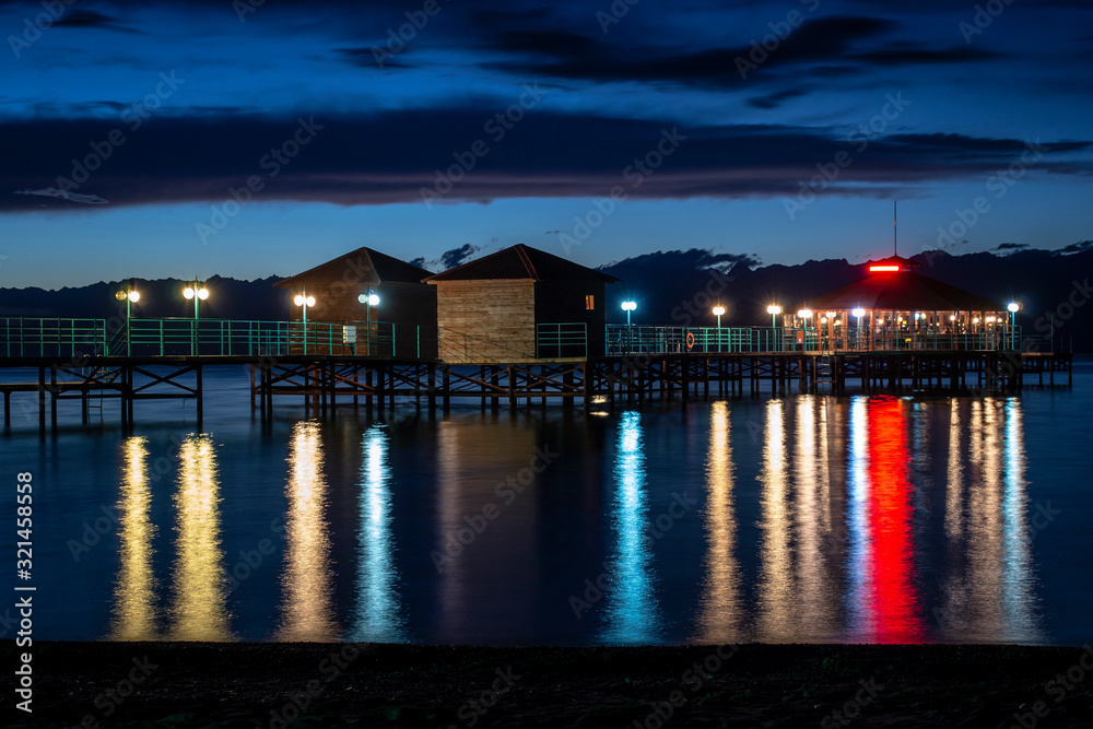 Night pier with colorful reflections on water and cloudy sky background.
