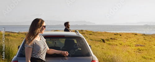 A man and a woman stand on the coast near the car.  Rest near the sea. Euro-trip. Heterosexual couple during a summer car trip. Panoramic coastal landscape with people.