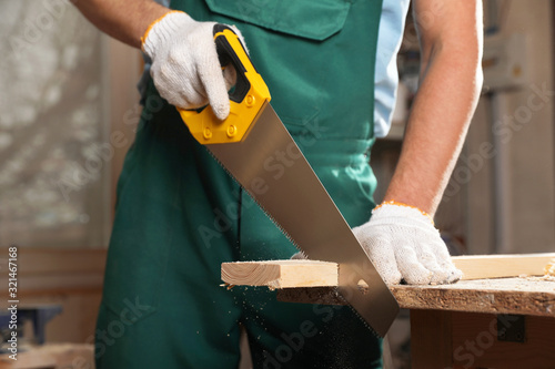 Professional carpenter cutting wooden board with handsaw in workshop, closeup photo