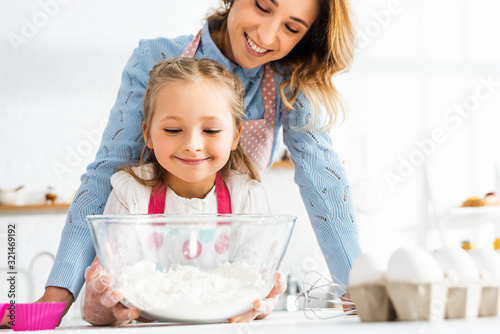 Selective focus of smiling mother and daughter near bowl with flour at kitchen table