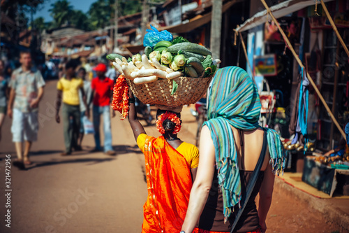 Quadro su tela An Indian woman in red Sari carries a large basket of vegetables on her head
