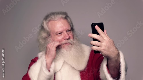 Senior Caucasian man in Santa Claus suit with splendid grey hair, mustache and beard, twirling moustache and looking at sell phone. Funny portrait of handsome European merry people on white background