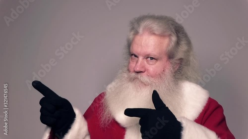 Senior Caucasian smiling man in Santa Claus suit with splendid grey hair, mustache and beard is pointing fingers at something and giving a high value. Funny portrait of handsome European people.