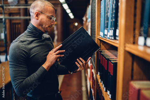 Serious male student standing near bookshelves checking literature for researching information, concentrated smart historian holding book in public library working with scientist publication photo