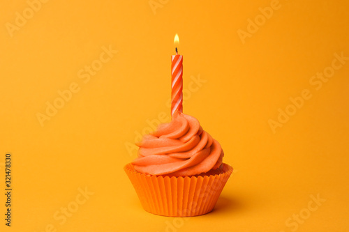 Delicious birthday cupcake with orange cream and burning candle on yellow background