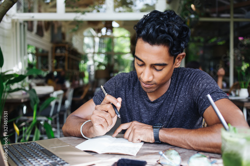 Thoughtful adult ethnic man reading notepad while sitting at laptop in cafe