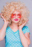 Beautiful retro-style blonde girl with voluminous curly hairstyle, in a blue polka-dot blouse and pink glasses on a gray background