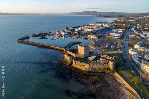 Medieval Norman Castle in Carrickfergus and Belfast Lough in sunrise light. Aerial view with marina, yachts, parking, breakwater, groyne, sediments and far view of Belfast in the background