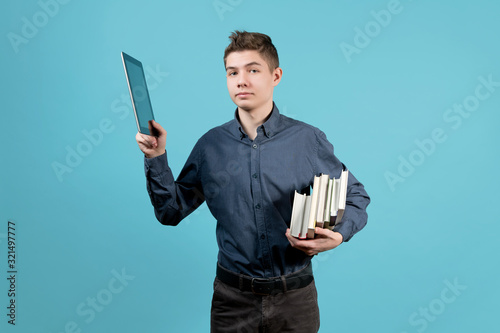 A teenager in a blue shirt holds a tablet and a stack of books under his armpit. Raises the tablet and looks pointedly at the camera photo