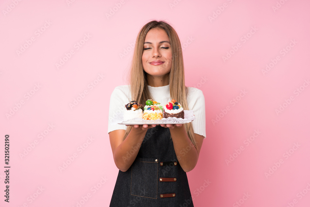 Young blonde woman over isolated pink background holding mini cakes enjoying the smell of them Young blonde woman over isolated pink background holding mini cakes enjoying the smell of them
