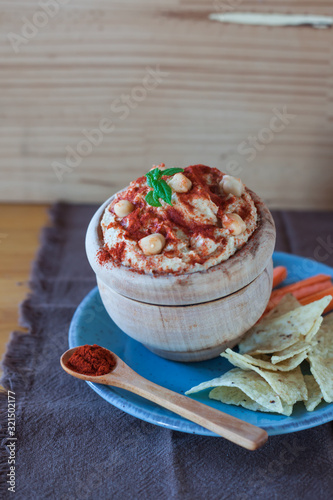 Wooden bowl with chickpea hummus and red paprika on a blue plate with carrot sticks and nachos with a rustic background of a brown and wooden handkerchief photo