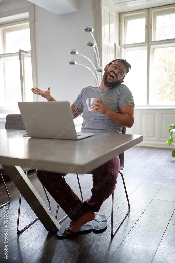 Young handsome man with beard and glasses, smiling an talking on cell phone while sitting at the table and laptop on it. modern, casual, business concept Young handsome man with beard and glasses, smiling an talking on cell phone while sitting at the table and laptop on it. modern, casual, business concept