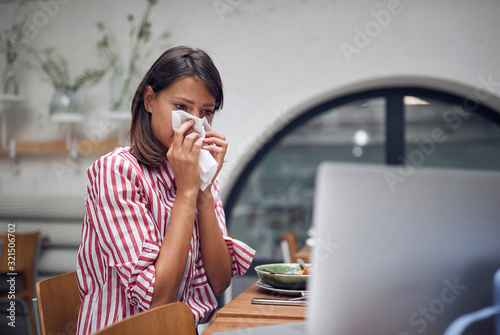 sick woman at caffe working on laptop. Blowing nose on tissue.