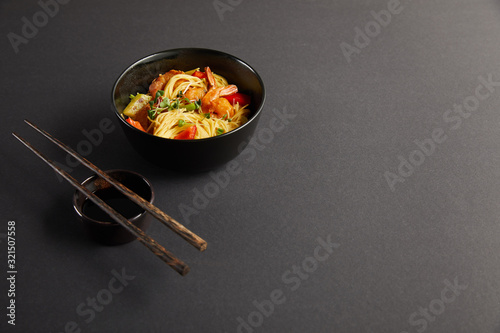 noodles with shrimps and vegetables in bowl near wooden chopsticks and soy sauce on black background