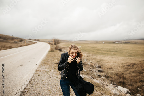 Portrait of young woman with hair blowing in the wind. She is  wearing a leather jacket with a blurred nature and road in background. Wanderlust autumn travel, atmospheric moment. 