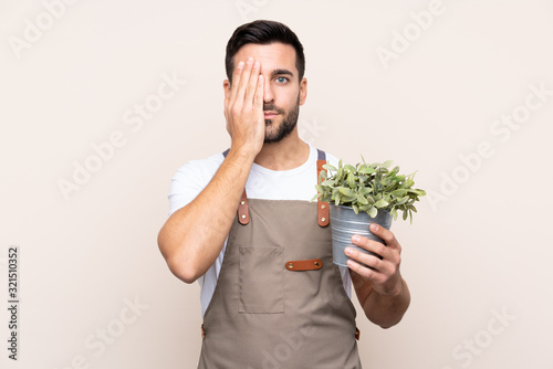 Gardener man holding a plant over isolated background covering a eye by hand