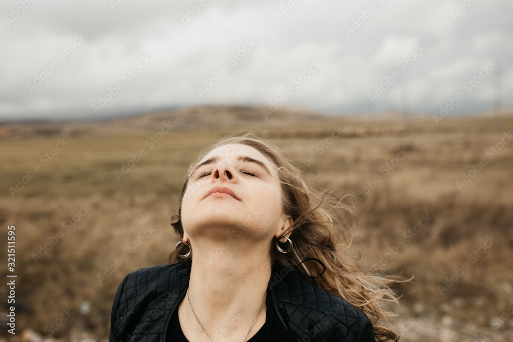Portrait of young woman with hair blowing in the wind. She is wearing a leather jacket with a blurred nature and road in background. Wanderlust autumn travel, atmospheric moment. Portrait of young woman with hair blowing in the wind. She is wearing a leather jacket with a blurred nature and road in background. Wanderlust autumn travel, atmospheric moment.