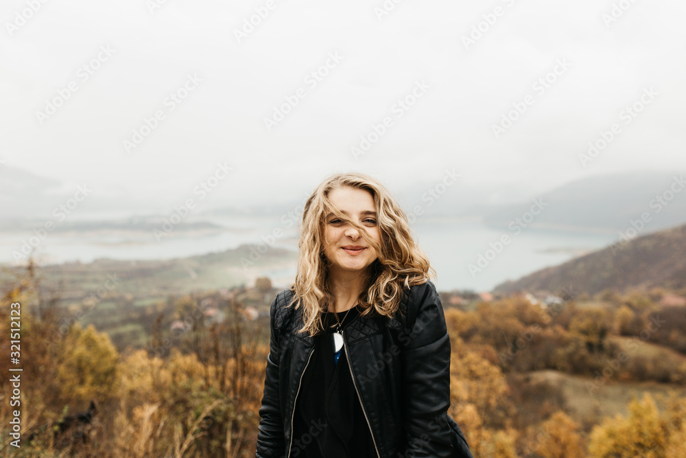 Portrait of young woman with hair blowing in the wind. She is  wearing a leather jacket with a blurred nature and road in background. Wanderlust autumn travel, atmospheric moment. 