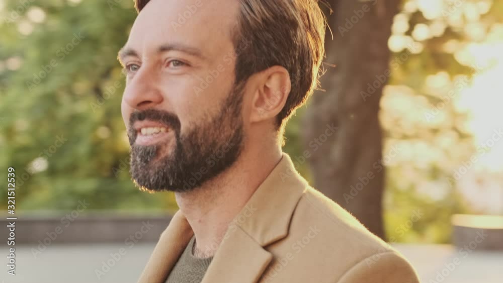 Smiling elegant bearded man in coat looking away while walking in the park outdoors