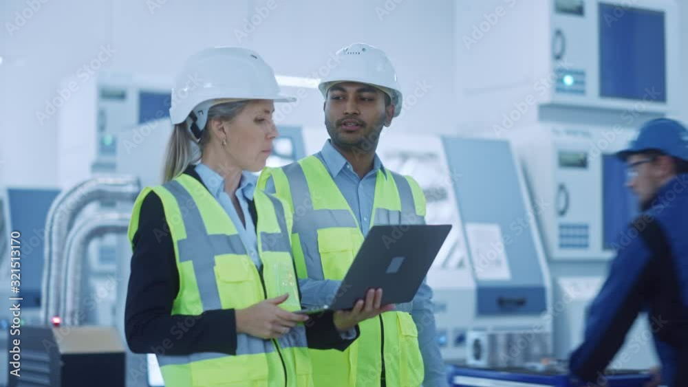 Chief Engineer and Project Manager Wearing Safety Vests and Hard Hats Walk Through Modern Factory, Talking, Optimizing Production Line. Industrial Facility: Professionals Working on Machinery Chief Engineer and Project Manager Wearing Safety Vests and Hard Hats Walk Through Modern Factory, Talking, Optimizing Production Line. Industrial Facility: Professionals Working on Machinery