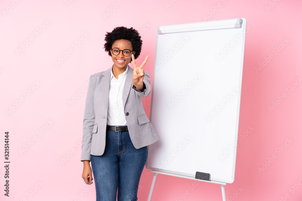 African american business woman giving a presentation on white board over isolated pink background smiling and showing victory sign African american business woman giving a presentation on white board over isolated pink background smiling and showing victory sign