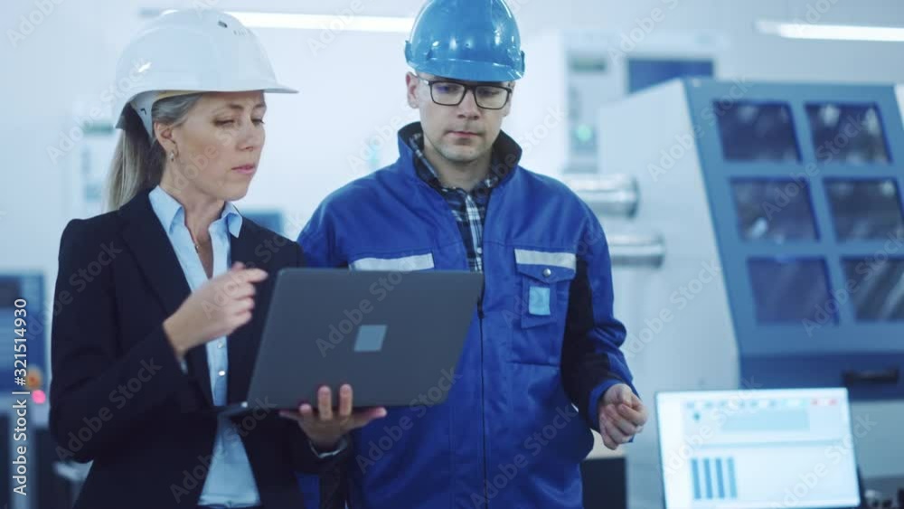 Female Manager and Project Engineer Wearing Hardhats Walk Through Industrial Factory, Use Laptop, Talk, Plan Productivity Optimization. Production Line Workers Operate CNC Machinery, Program robot arm Female Manager and Project Engineer Wearing Hardhats Walk Through Industrial Factory, Use Laptop, Talk, Plan Productivity Optimization. Production Line Workers Operate CNC Machinery, Program robot arm