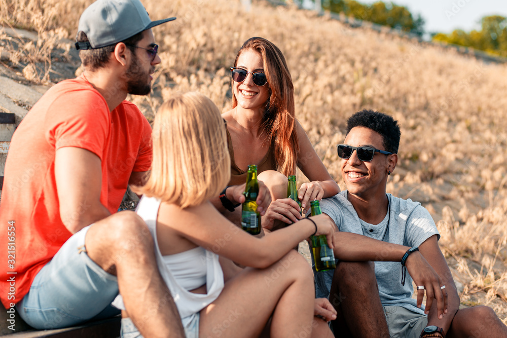 Group of friends having fun, talking and drinking beer while sitting on the stairs of riverbank.