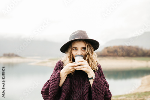 Happy beautiful woman with hat drinking coffee in nature, on a foggy morning. Lake and mountains are in the background. 