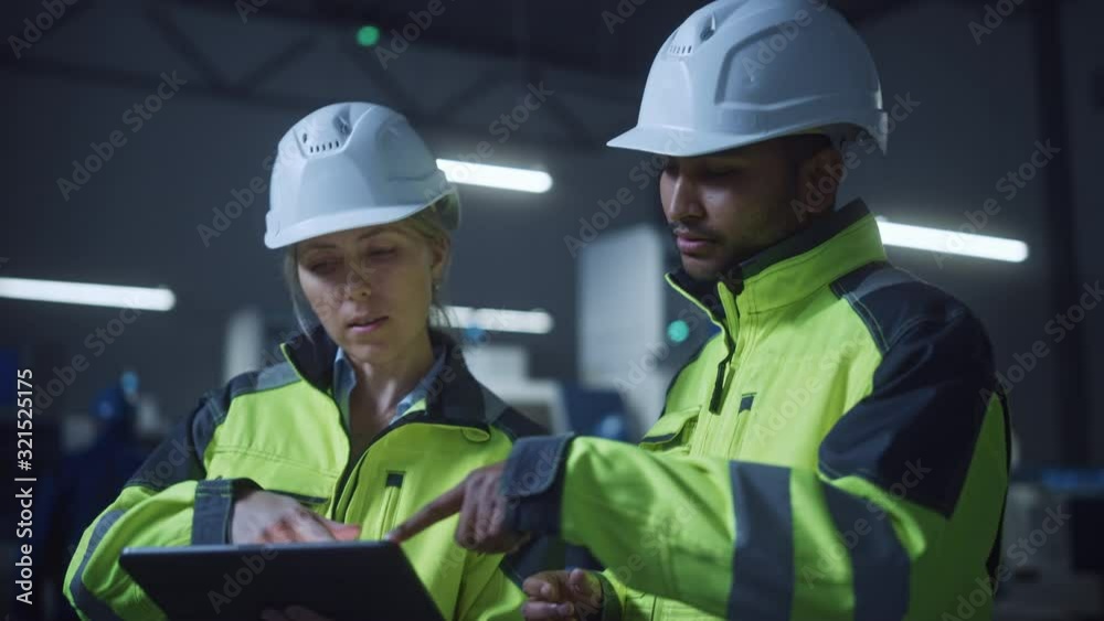 Chief Engineer and Project Manager Wearing Safety Vests and Hard Hats, Use Digital Tablet Computer in Modern Factory, Talking, Optimizing Production Line. Industrial Facility with CNC Machinery