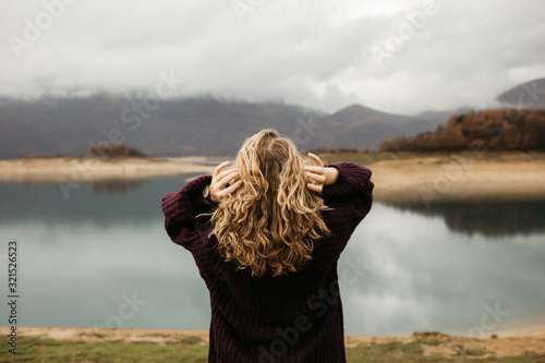 happy girl with curly blond hair dances on a lake alone, her hair is flying because of the wind flow, free as a bird. photo of girl with curly hair standing back to camera over lake background.