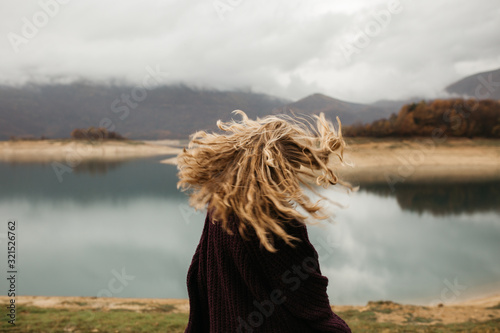 happy girl with curly blond hair dances on a lake alone, her hair is flying because of the wind flow, free as a bird. photo of girl with curly hair standing back to camera over lake background.