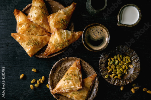 Homemade Turkish traditional dessert baklava with pistachio served on ceramic plate with bowl of nuts, cup of coffee, cream, cezva over black wooden background. Flat lay, space