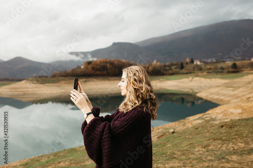 Beautiful woman with curly hair holding mobile phone and taking photos of lake on a cloudy day. She is texting on smartphone and taking selfie. 