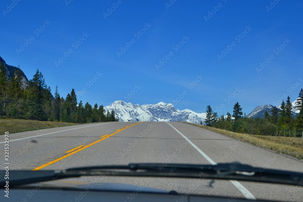 Road from behind the window screen of the car. Gray road lined with forest, high mountains covered with snow, blue sky. Canadian Rockies.