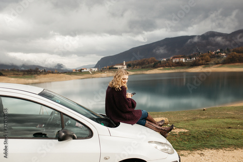 Beautiful woman with curly hair, sitting on the car, holding mobile phone and taking photos of lake on a cloudy day. She is texting on smartphone and taking selfie.  © Melika