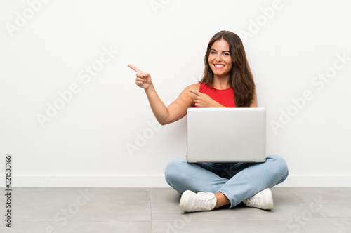 Young woman with a laptop sitting on the floor pointing finger to the side