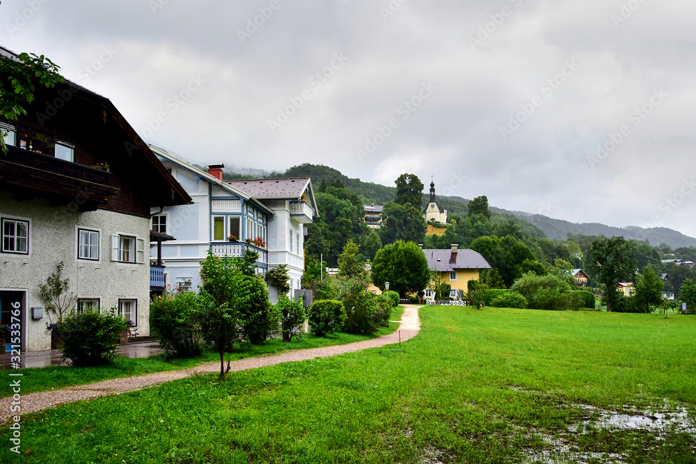 Mondsee, Austria - A row of houses in the countryside, next to a green lawn, in the background you can see a church standing on a hill among green trees, in the summer afternoon.