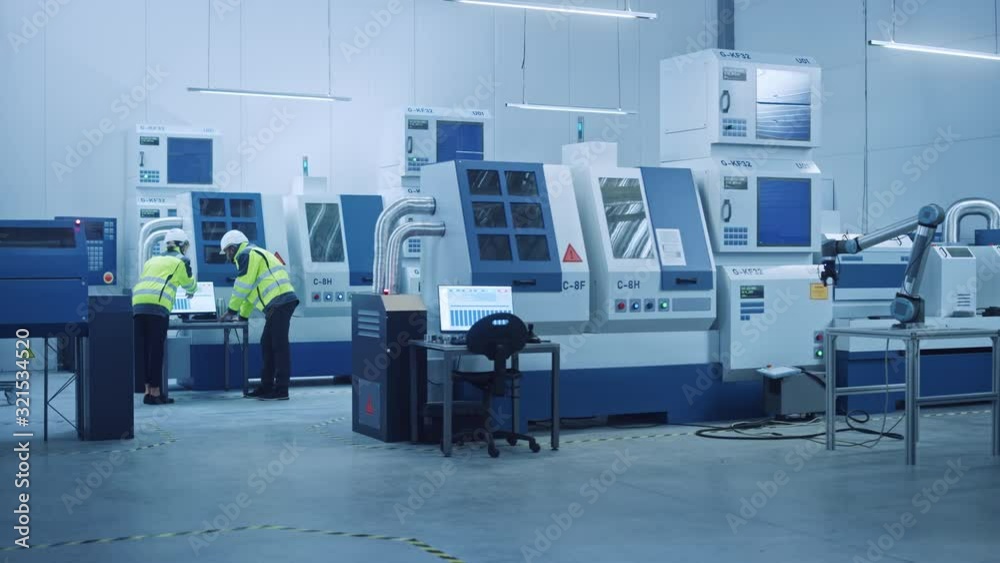 Chief Engineer and Project Manager Wearing Safety Vests and Hard Hats Work on Compute, Talk, Optimize Production Line. On Foreground: industry 4.0 Modern Factory with CNC Machinery and robot arm Chief Engineer and Project Manager Wearing Safety Vests and Hard Hats Work on Compute, Talk, Optimize Production Line. On Foreground: industry 4.0 Modern Factory with CNC Machinery and robot arm
