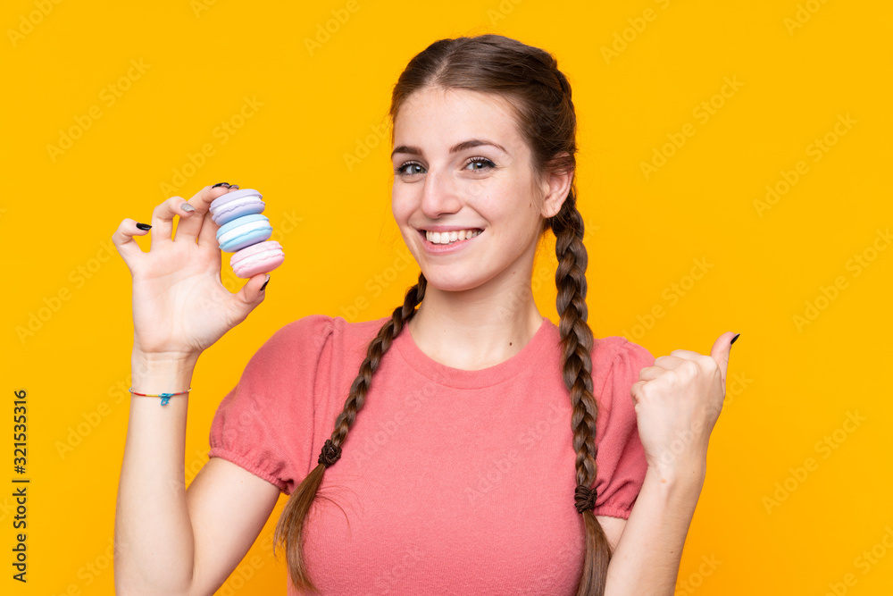 Young woman over isolated yellow background holding colorful French macarons and pointing side Young woman over isolated yellow background holding colorful French macarons and pointing side