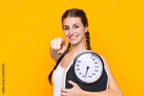 Young woman over isolated yellow background holding a weighing machine and pointing to the front