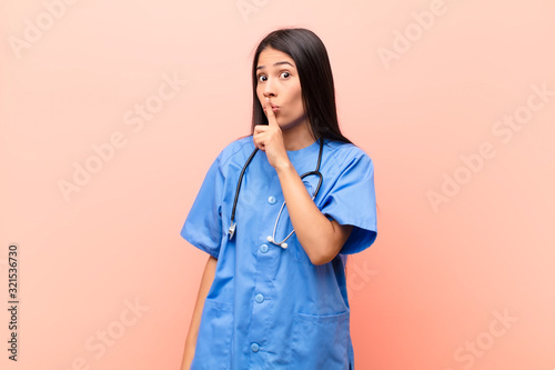 young latin nurse asking for silence and quiet, gesturing with finger in front of mouth, saying shh or keeping a secret against pink wall