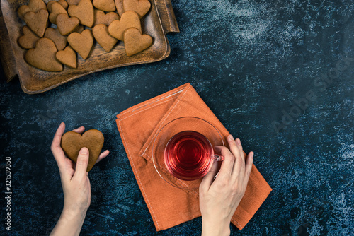 Homemade baked ginger cookies lie on a wooden board. Shape in the form of hearts. A loved breakfast with a red tea on a holiday or an evening party of Valentines Day. Dark flat lay with female hands.