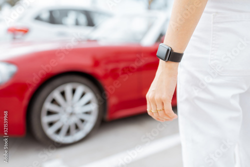 Woman with smartwatch at the car parking with red vehicle on the background, close-up view on watch with black screen. Car control with a smart watch concept