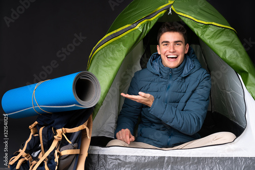 Teenager caucasian man inside a camping green tent isolated on black background presenting an idea while looking smiling towards