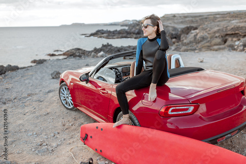 Portrait of a young woman surfer in swimsuit sitting with surfboard on the red cabriolet on the rocky ocean coast. Active lifestyle and surfing concept photo